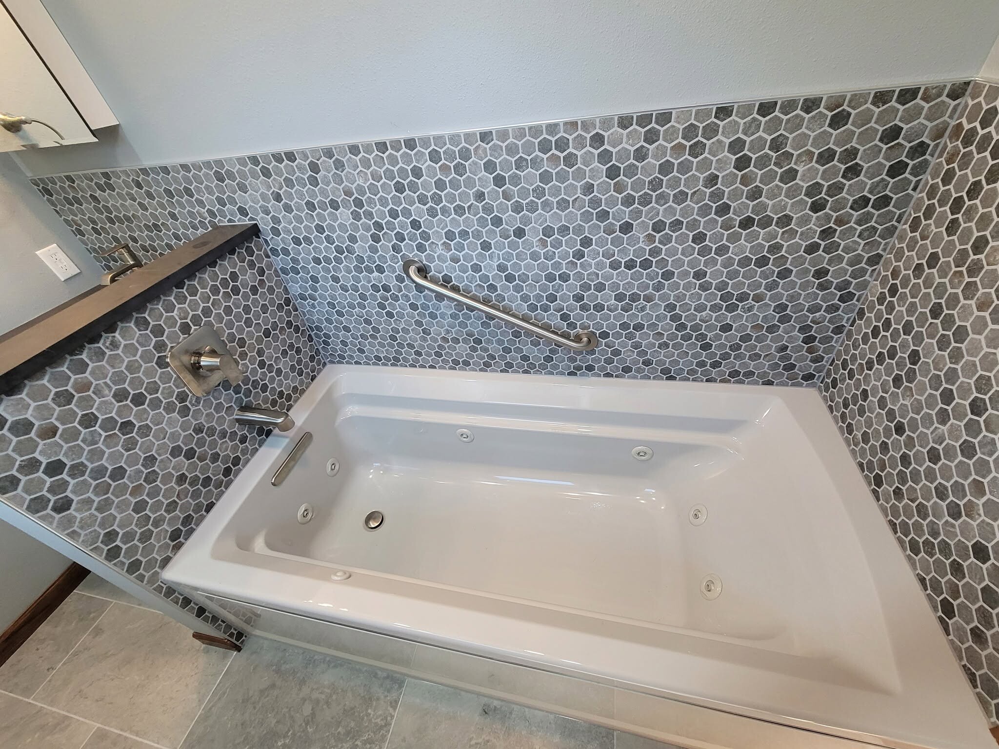 Finished jetted tub set in a hexagon mosaic surround with a brushed nickel grab bar and a solid surface deck cap