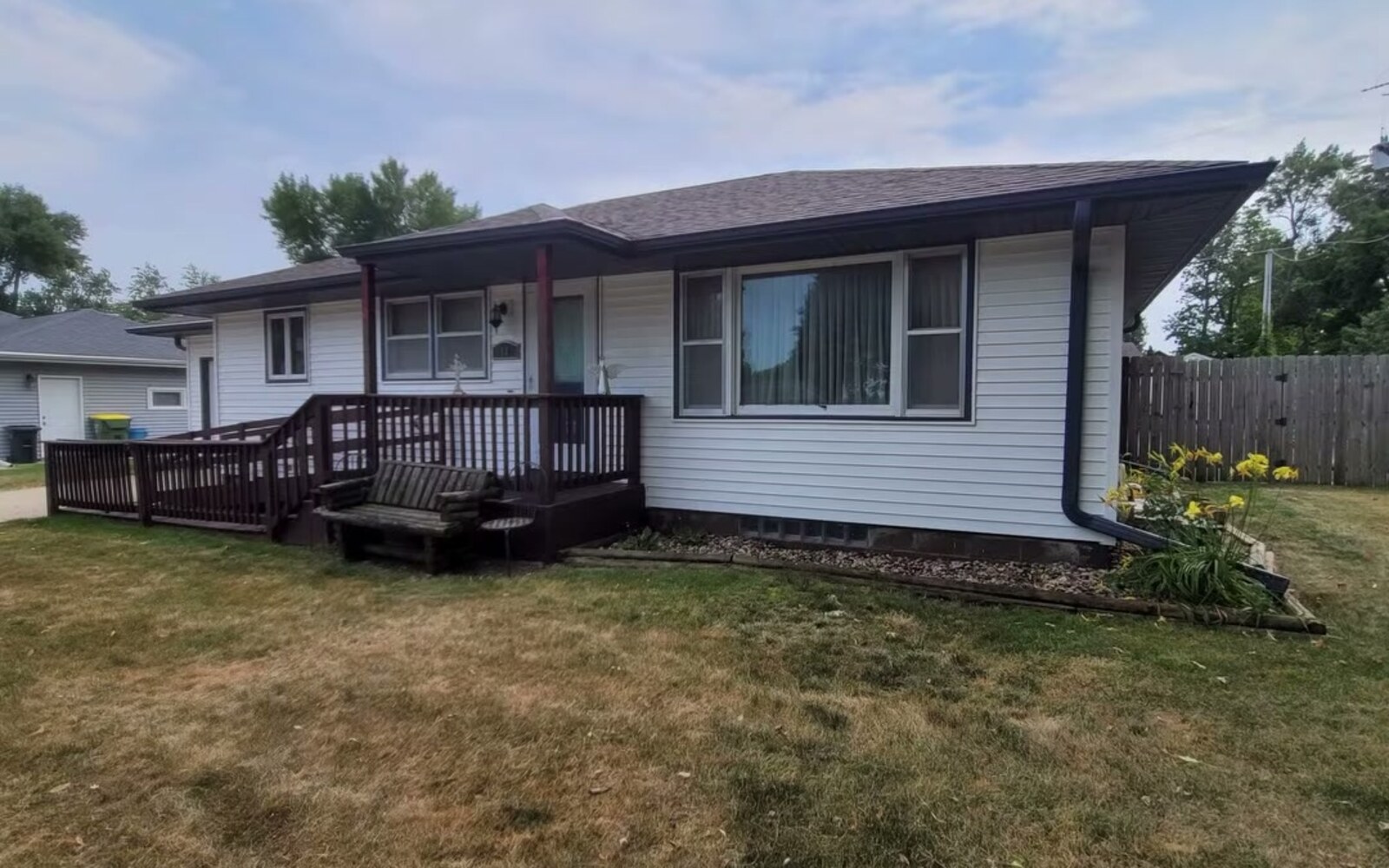 Washed white house siding with a dark stained deck ramp and a rock bed of yellow daylilies along the foundation